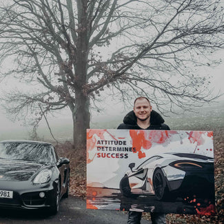 Mann hält Motivationsbild mit Sportwagen und Aufschrift 'Attitude determines success', daneben schwarzes Auto in nebliger Landschaft.
