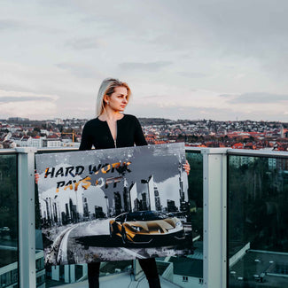 Frau hält Motivationsbild mit Sportwagen und Skyline, Text 'Hard work pays off', auf Balkon mit Stadtblick.